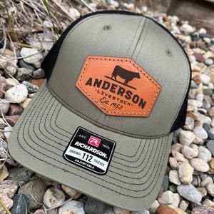 May include: A green and black trucker hat with a brown leather patch that says "Anderson Livestock Est. 1953" and a black silhouette of a cow. The hat also has a red and white label that says "Richardson 112 Trucker Adjustable Est. 1970".