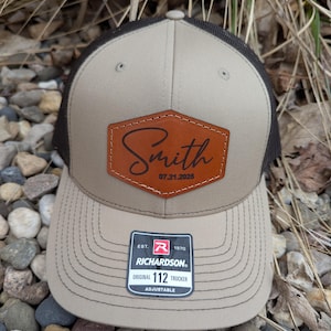 May include: A tan and black trucker hat with a brown leather patch that says "Smith 07.21.2025". The hat has a black mesh back and a tan front with a black "Richardson" label that says "Est. 1970 Original 112 Trucker Adjustable".