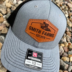 May include: A gray and black trucker hat with a brown leather patch that says "Smith Farms Est. 1970". The hat also has a red and white label that says "Richardson 112 Trucker Adjustable".