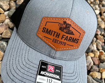 Peut inclure: Casquette de camionneur grise et noire avec un écusson en cuir marron portant l'inscription "SMITH FARMS EST.1970" et un dessin de moissonneuse-batteuse. La casquette a une visière incurvée et un dos en filet. L'étiquette indique "RICHARDSON ORIGINAL 112 TRUCKER".