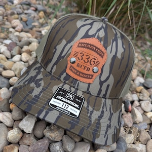 May include: A camouflage trucker hat with a brown leather patch. The patch reads "Battalion Chief 336 KCFD Fire & Rescue". The hat has a mesh back and a Richardson 112 Trucker label.