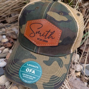 May include: A camouflage green and brown trucker hat with a brown leather patch that says "Smith 07.21.2025". The hat has a mesh back and a green sticker that says "Legacy. OFA Visor Board Made From Recycled Content Live Your Legacy."