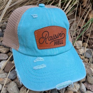 May include: A turquoise blue and brown distressed trucker hat with a brown leather patch that says "Raisin' Hell" in a cursive font.
