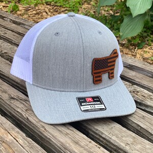 May include: Gray and white trucker hat with a brown leather patch of a steer with an American flag design. The patch is on the front of the hat. The hat has a white mesh back and a black label with the text "Richardson. Original Trucker 112 Adjustable" on the back.
