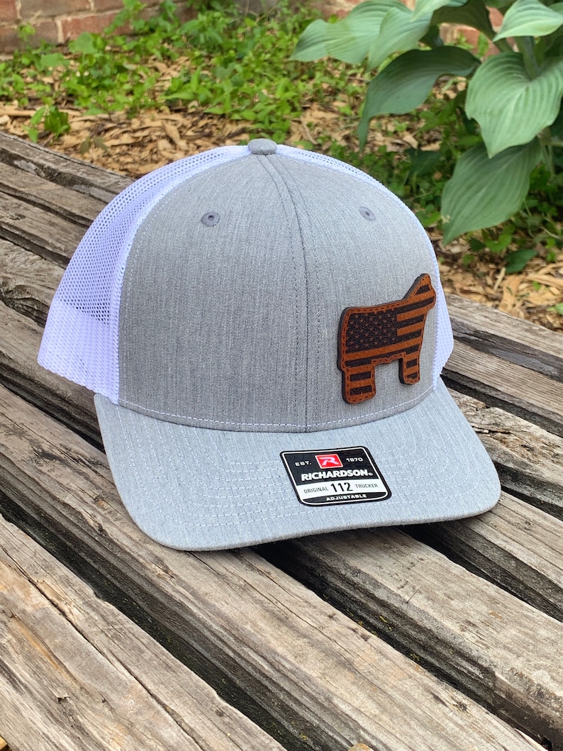 May include: Gray and white trucker hat with a brown leather patch of a steer with an American flag design. The patch is on the front of the hat. The hat has a white mesh back and a black label with the text "Richardson. Original Trucker 112 Adjustable" on the back.