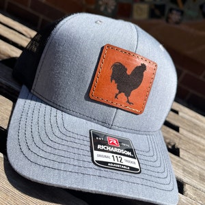 May include: A gray and black trucker hat with a brown leather patch featuring a rooster silhouette. The patch has stitching around the edges. The hat has a black mesh back and a black adjustable strap with a black label that reads "EST. 1970 RICHARDSON. ORIGINAL 112 TRUCKER ADJUSTABLE".