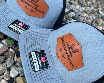 May include: Two gray trucker hats with black mesh backs. Each hat features a brown leather patch with the text "Salsbury Ranch Est. 2011" and a graphic of an animal. The hats have a curved brim and an adjustable strap.