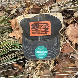 May include: A black and brown trucker hat with a brown leather patch featuring an American flag and a silhouette of a lineman working on a power line. The hat has a mesh back and a green sticker that says "Old Favorite Trucker Legacy Premium Vintage Inspired Caps for a Relaxed Lifestyle #OldFavoriteTrucker #Legacy".