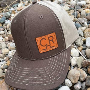 May include: A brown and beige baseball cap with a brown leather patch that says "CR" in orange letters.