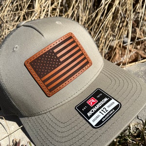 May include: A green and black trucker hat with a brown leather American flag patch and a black and white label that reads "EST. 1970 RICHARDSON® ORIGINAL 112 TRUCKER ADJUSTABLE".
