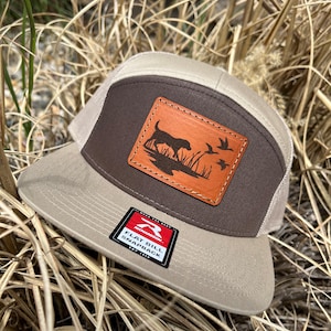 May include: A brown and beige flat bill snapback hat with a leather patch featuring a dog and ducks. The patch is stitched onto the hat. The hat has a Richardson logo on the front.