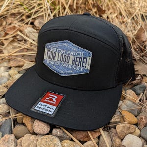 May include: Black flat bill snapback hat with a black mesh back. The hat has a custom patch with the text "CUSTOM PATCH YOUR LOGO HERE! SHAPES AVAILABLE".