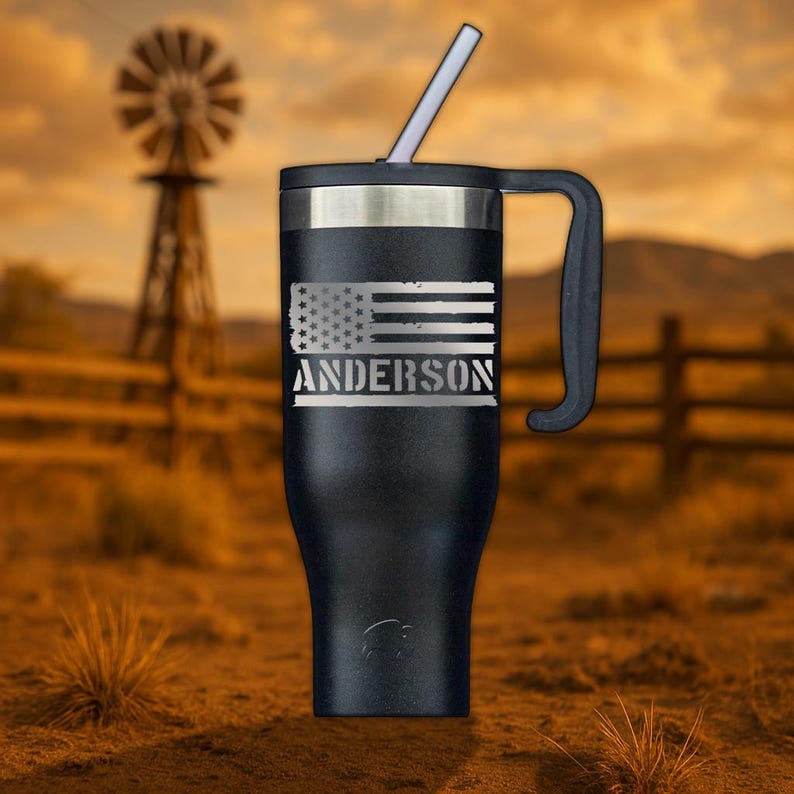 May include: Black stainless steel travel mug with a handle and a straw. The mug has a distressed American flag design with the name "ANDERSON" engraved below it.