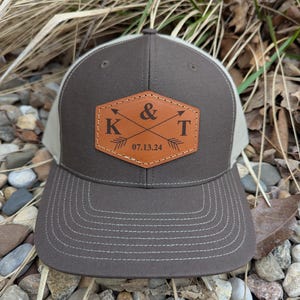 May include: A brown and beige trucker hat with a brown leather patch that says "K & T" with crossed arrows and the date "07.13.24".