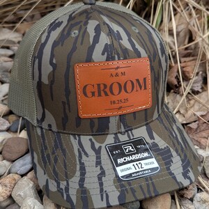 May include: A camouflage trucker hat with a brown leather patch that says "A&M Groom 10.25.25" and a black and white label that says "Richardson 112 Original Trucker Adjustable".