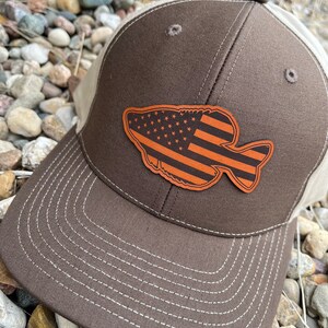 May include: A brown and beige baseball cap with a brown leather patch in the shape of a fish with an American flag design.