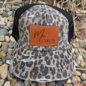 May include: A black and brown leopard print trucker hat with a brown leather patch that says "Mrs. Smith" in cursive.