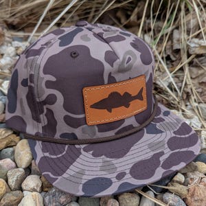 May include: A camouflage baseball cap with a brown leather patch featuring a silhouette of a fish. The cap has a brown rope band.