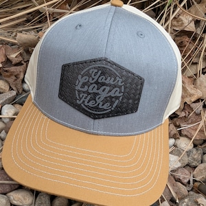 May include: A gray and beige baseball cap with a brown leather patch that says "Your Logo Here!" The patch has a woven texture.