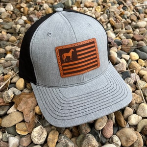 May include: A gray and black baseball cap with a brown leather patch featuring a silhouette of a horse and an American flag design.