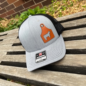 May include: Gray and black trucker hat with a brown leather cow tag on the front. The tag has a black silhouette of a cow. The hat has a black mesh back and a black brim. The hat has a white label on the side that says "Est. 1970 Richardson. Original Trucker 112 Adjustable."