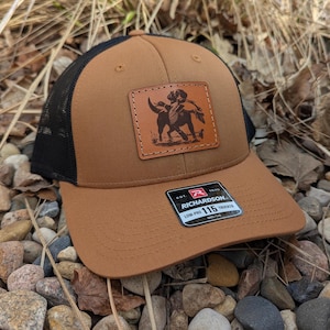 May include: A brown and black trucker hat with a leather patch featuring a dog and a duck. The patch is labeled "Richardson 115 Trucker".