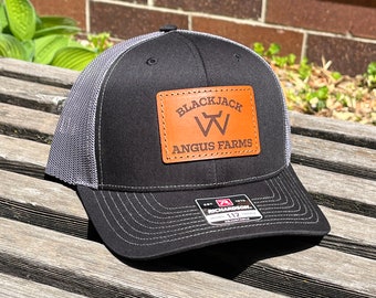 May include: Black and gray trucker hat with a leather patch. The patch features the words "Blackjack Angus Farms" and a stylized "W" logo. The hat has a curved brim and a mesh back.