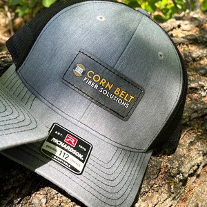 May include: Gray and black trucker hat with a black patch that says "CORN BELT FIBER SOLUTIONS". The hat also has a red and white patch that says "RICHARDSON. EST. 1970 ORIGINAL 112 TRUCKER ADJUSTABLE".
