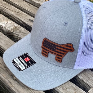 May include: A gray and white trucker hat with a brown leather American flag steer head patch. The hat has a mesh back and an adjustable strap. The hat is made by Richardson 112.