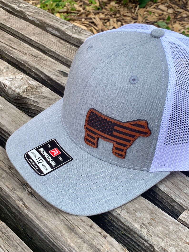 May include: A gray and white trucker hat with a brown leather American flag steer head patch. The hat has a mesh back and an adjustable strap. The hat is made by Richardson 112.