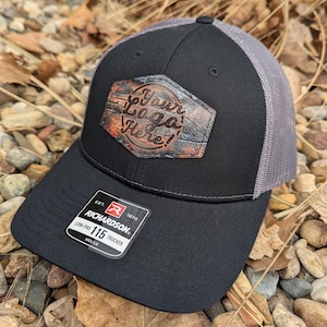 May include: Black and gray trucker hat with a brown leather patch that says "Your Logo Here!" The hat is made by Richardson and has a low-profile fit.