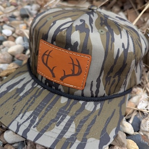 May include: A camouflage baseball cap with a brown leather patch featuring a deer antler design.