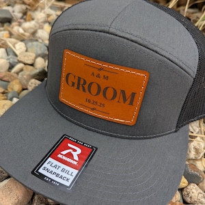 May include: A gray and black trucker hat with a brown leather patch that says "A&M GROOM 10.25.25". The hat has a flat bill and a mesh back. The hat is made by Richardson.