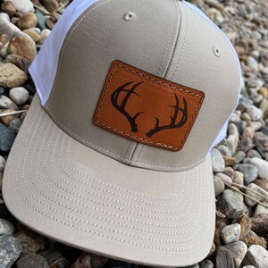 May include: A beige and white trucker hat with a brown leather patch featuring a deer antler design.