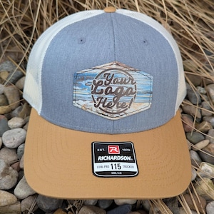 May include: A gray and white trucker hat with a brown leather patch that says "Your Logo Here!" The hat has a tan brim and a Richardson 115 logo on the back.