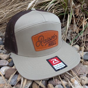 May include: A tan and brown flat bill baseball cap with a brown leather patch that says "Raisin' Hell" in cursive. The cap has a mesh back and a red and white logo that says "Rich Dad" on the side.