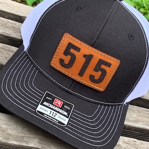 May include: Black and white trucker hat with a brown leather patch that says "515". The hat has a white mesh back and a black front. The hat is adjustable and has a Richardson 112 label.