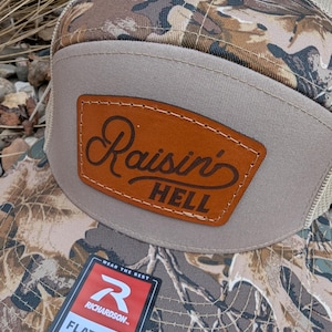 May include: A brown and tan camouflage trucker hat with a brown leather patch that says "Raisin' Hell". The hat has a mesh back and a flat bill. The hat also has a red and white label that says "Richardson" and "Flat Bill Snapback Est 1970".