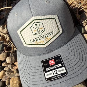 May include: A gray trucker hat with a black mesh back. The hat features a patch with the text "LAKEVIEW Country Club" and golf-related graphics. A black and white tag with the Richardson logo is attached.