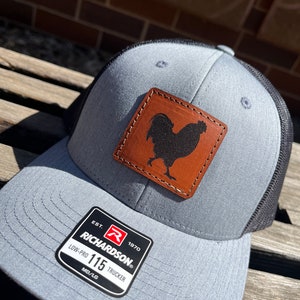 May include: A gray and black trucker hat with a brown leather patch featuring a rooster silhouette. The patch has the text "Richardson" and "Est. 1970". The hat has a label that says "Low-Pro 115 Trucker" and "MD/LG".