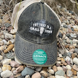 May include: A black and beige trucker hat with a mesh back. The hat has a black patch with glitter and the text "TRY THAT IN A SMALL TOWN" with a skull graphic. The hat also has a green and white sticker with the text "OLD FAVORITE TRUCKER LEGACY PREMIUM, VINTAGE-INSPIRED CAPS FOR A RELAXED LIFESTYLE #LIVETOURLEGACY"