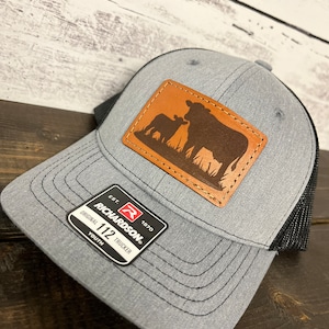 May include: Gray and black youth trucker hat with a brown leather patch featuring a silhouette of a cow and calf. The hat has a Richardson 112 logo on the side.