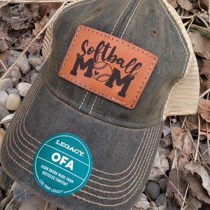 May include: A green and brown baseball cap with a brown leather patch that says "Softball Mom" with a baseball graphic. The cap has a mesh back and a green sticker that says "Legacy OFA Visor Board Made From Recycled Content Live Your Legacy".