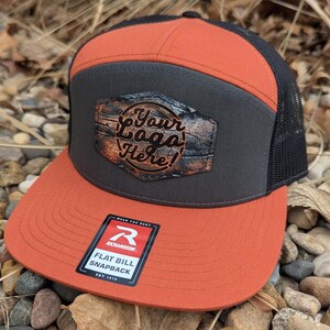 May include: A black and orange flat bill snapback hat with a brown woodgrain patch that says "Your Logo Here!" and a black mesh back. The hat has a Richardson logo on the side.
