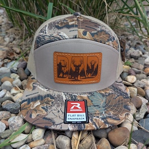 May include: A flat bill snapback hat with a camouflage pattern on the bill and top. The hat features a tan front panel with a brown leather patch displaying a deer and hunter silhouette. The Richardson brand tag is visible.