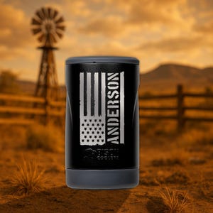 May include: A black and silver personalized can cooler with an American flag design and the name "ANDERSON" engraved on the side. The cooler is sitting on a brown surface with a windmill in the background.