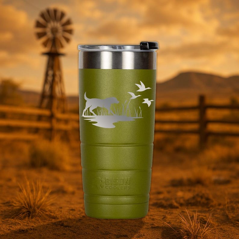 Puede incluir: Un vaso de acero inoxidable verde con un dise&ntilde;o grabado con l&aacute;ser de un perro corriendo por un campo con p&aacute;jaros volando por encima. El vaso tiene una tapa negra y el texto "Bison Coolers" est&aacute; grabado en la parte inferior.