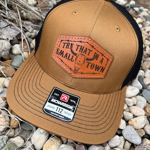 May include: A brown and black trucker hat with the text "TRY THAT IN A SMALL TOWN" on a leather patch. The hat has a curved brim and a mesh back. The Richardson brand label is visible.