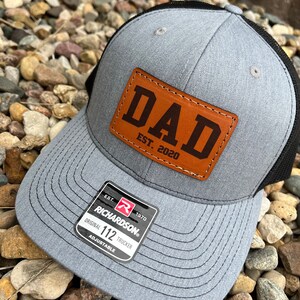 May include: Gray trucker hat with a black mesh back and a brown leather patch that reads "DAD EST. 2020." The hat has a curved brim and an adjustable strap. A Richardson 112 Trucker tag is visible.