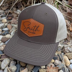 May include: A brown and beige baseball cap with a brown leather patch that says "Smith 07.21.2025" on the front.
